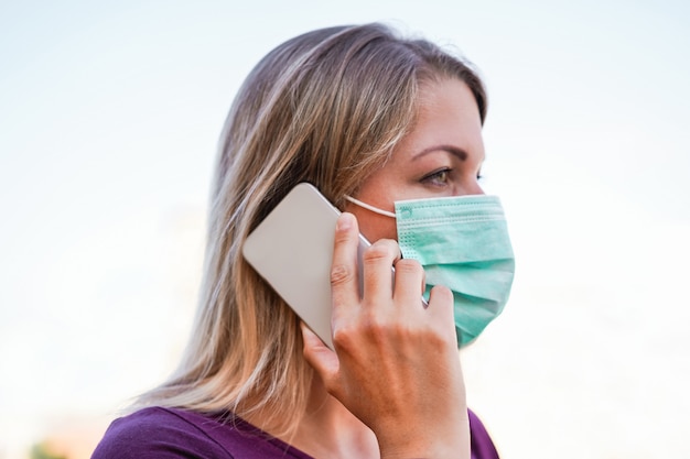 Junge Frau, die mit Handy im Freien trägt Gesichtsmaske spricht