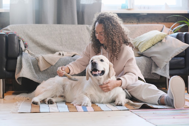 Junge Frau, die mit ihrem Hund auf dem Boden sitzt und sein Fell im Wohnzimmer zu Hause kämmt