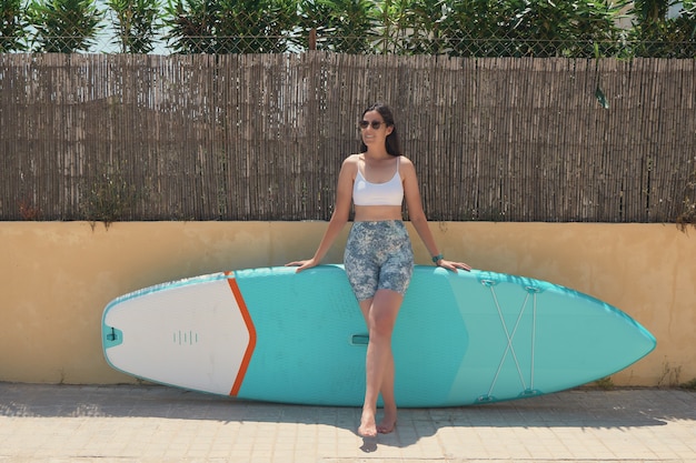 Junge Frau, die mit ihrem Paddel-Surfbrett an einer Wand lächelt