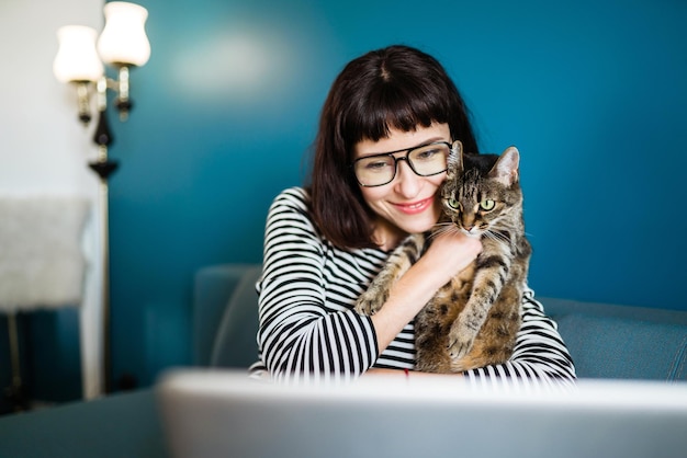 Junge Frau, die mit Laptop arbeitet und mit einer Katze, die zu Hause im modernen Wohnzimmer sitzt