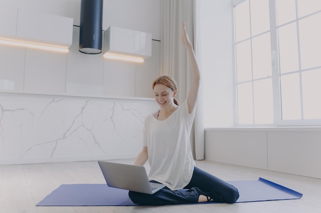 Junge Frau, die mit Laptop auf den Knien auf dem Boden sitzt Online-Fitness-Tutorial Ferntraining