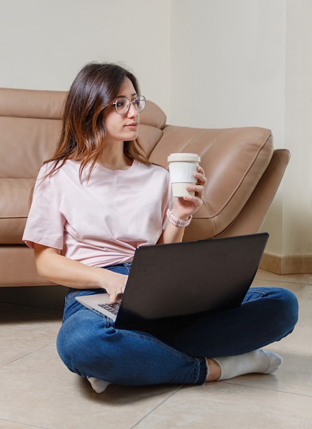 Junge Frau, die mit Laptop mit Kaffee arbeitet, um in Hand zu gehen, während auf dem Boden zu Hause sitzen
