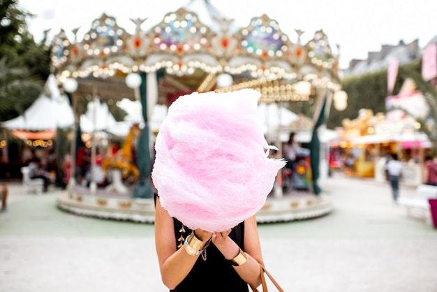 Junge Frau, die mit rosa Zuckerwatte draußen vor dem Karussell im Vergnügungspark steht