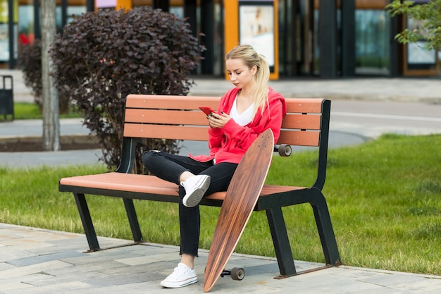 Junge Frau, die mit Skateboard oder Longboard auf Holzbank sitzt