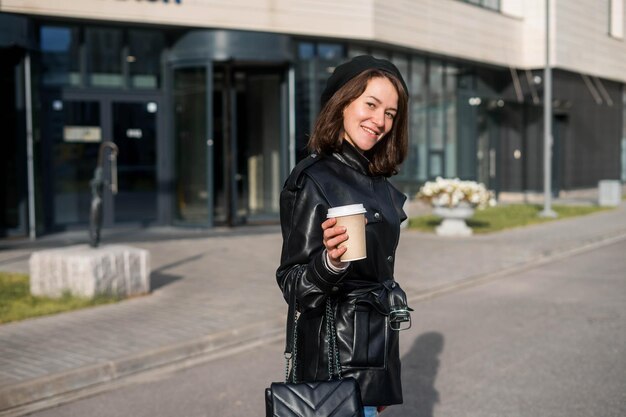 Junge Frau, die mit Smartphone und Kaffee vor dem Büro bleibt.