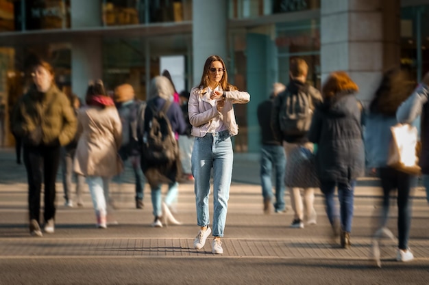 Junge frau, die mitten auf einer überfüllten straße spazieren geht und die zeit zur hand sieht, beobachtet das leben in der großstadt