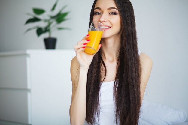 Junge Frau, die morgens ein Glas Orangensaft genießt, während sie sich entspannt, sitzend auf ihrem Bett in einem Konzept der gesunden Diät
