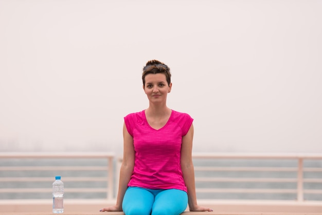 junge frau, die nach einem erfolgreichen trainingslauf auf der promenade am meer mit einer flasche wasser sitzt