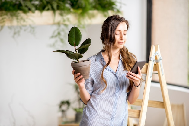 Junge Frau, die nach Hause mit Grün pflanzt, die mit Telefon und Blumentopf auf der Leiter steht