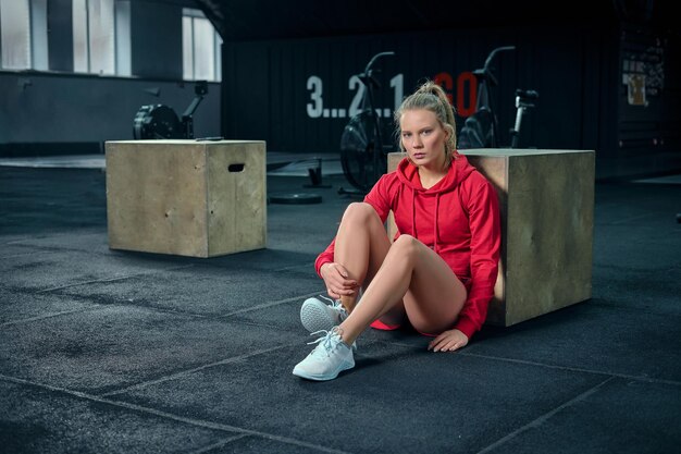 Junge Frau, die nach ihrem Training in der Nähe einer Kiste im Fitnessstudio sitzt und in die Kamera schaut