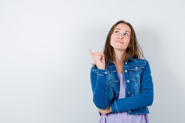 Junge Frau, die nach oben zeigt, im T-Shirt, in der Jacke nach oben schaut und verträumt aussieht. Vorderansicht.