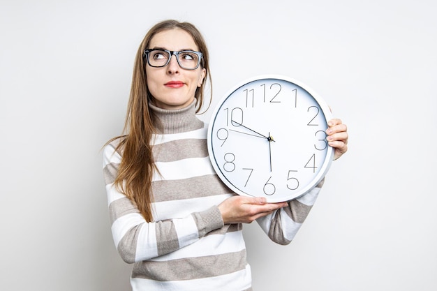 Junge Frau, die nachdenklich nach oben schaut und eine Uhr auf hellem Hintergrund hält