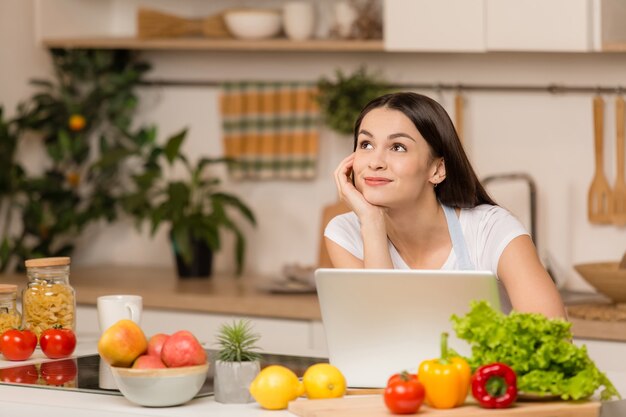 Junge Frau, die nahe Schreibtisch und Laptop in der Küche steht, lächelt, träumt. Food Blogger Konzept