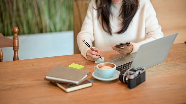 Junge Frau, die Notizen auf einem klebrigen bunten Briefpapier macht, während sie auf dem Smartphone schaut, sitzt im Café