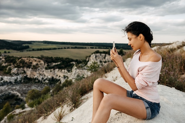 Junge Frau, die oben auf dem Berg sitzt und Bilder der Landschaft mit Smartphone macht.