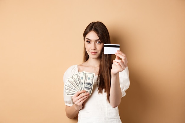 Junge Frau, die Plastikkreditkarte zeigt, bevorzugen kontaktlose Zahlung anstelle von Dollarnoten, die auf beigem Hintergrund stehen.