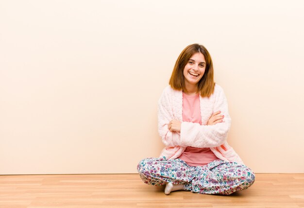 Junge Frau, die Pyjamas trägt, die zu Hause sitzen und wie ein glücklicher, stolzer und zufriedener Leistungsträger aussehen, der mit verschränkten Armen lächelt