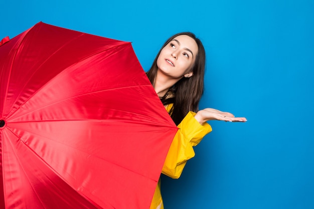 Junge Frau, die Regenmantel hält, der bunten Regenschirm über blauer Wand hält