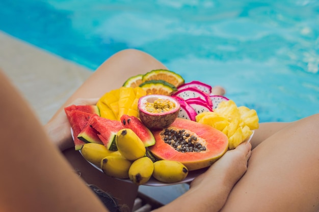 Junge Frau, die sich am Hotelpool entspannt und Obstteller isst. Exotische Sommerdiät. Foto von Beinen mit gesundem Essen am Pool, Draufsicht von oben. Tropischer Strandlebensstil