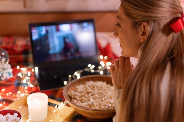 Junge Frau, die sich den Weihnachtsfilm auf dem Laptop mit Popcorn im Camping-Wohnwagen in einem gemütlichen Bett mit karierter und leuchtender Lichtergirlande nachts an Weihnachten oder Silvester ansieht. Hand aufs Gesicht