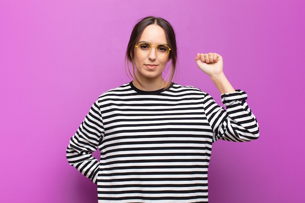 Junge Frau, die sich ernst, stark und rebellisch fühlt, Faust erhebt, protestiert oder für Revolution auf lila Wand kämpft