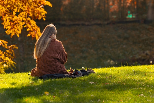 Junge Frau, die sich in einem Park im Herbst oder Herbst entspannt, sitzt auf einem grasbewachsenen Ufer mit Blick auf Wasser mit ihrem Rücken zur Kamera