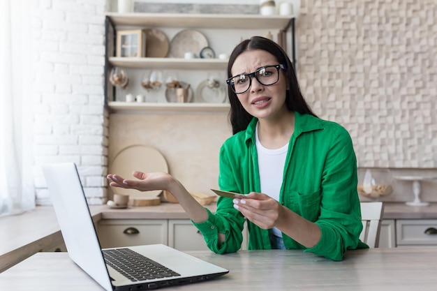 Junge Frau, die sich Sorgen um Kreditkartenprobleme macht, sitzt zu Hause in der Küche und hält einen Laptop in der Hand