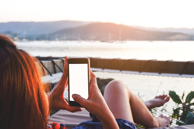 Junge Frau, die Smartphone mit leerem Bildschirm beim Lügen von Seitenmeer mit Abendsonnenuntergang verwendet.