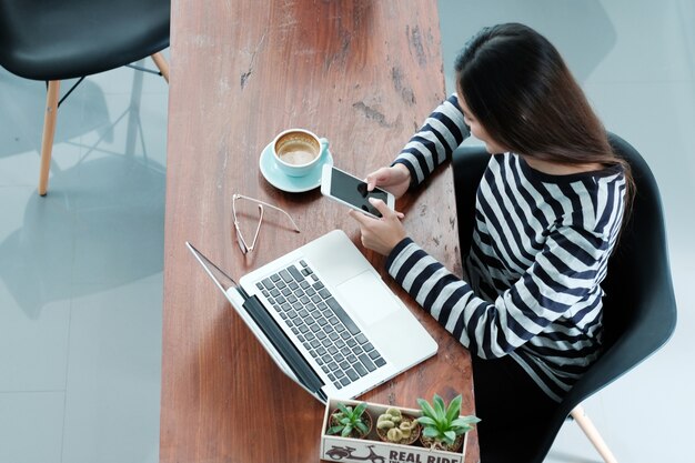 Junge Frau, die Smartphone und Schossspitzencomputer beim Sitzen am Caféhintergrund verwendet