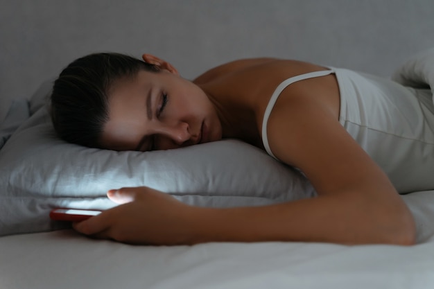 Junge Frau, die spät in der Nacht auf dem Bett mit Smartphone in ihrer Hand schläft