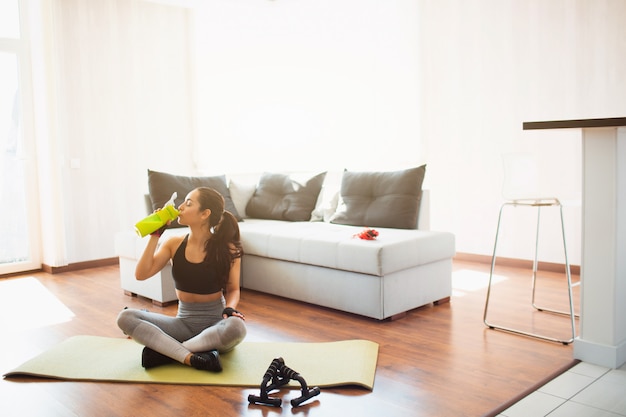 Junge Frau, die Sporttraining im Raum während der Quarantäne tut. Setzen Sie sich mit gekreuzten Beinen auf die Matte und trinken Sie Protein aus der grünen Flasche. Ruhe entspannen nach dem Training.