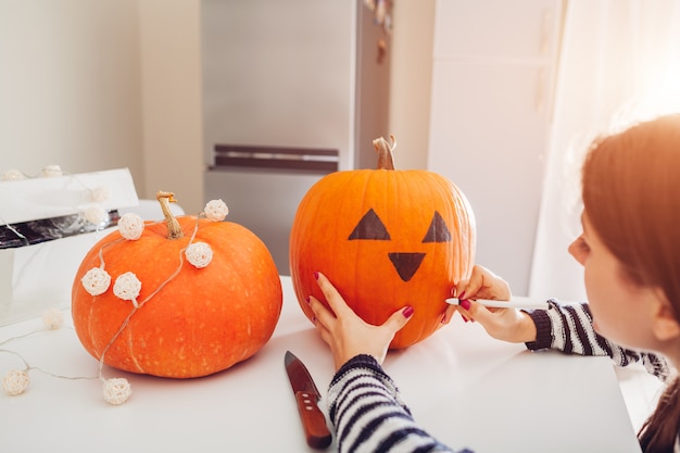 Junge Frau, die Steckfassung-Olaterne für Halloween auf Küche bildet. Zeichnen von Augen, Nase und Mund mit Stift auf Kürbis