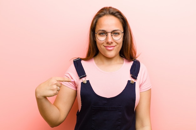 Junge Frau, die stolz, zuversichtlich und glücklich schaut, lächelt und auf sich selbst zeigt oder Nummer eins Zeichen gegen Rosa macht