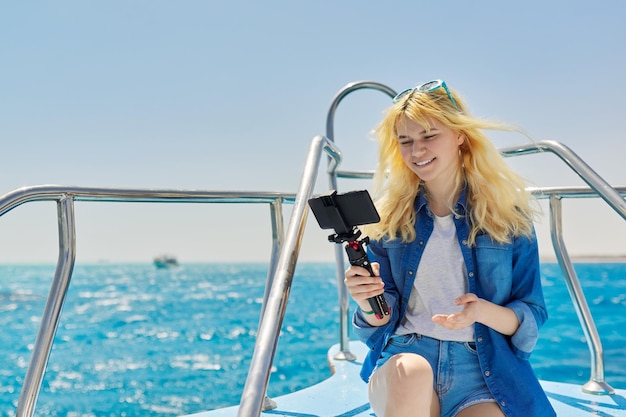 Junge Frau, die Stream auf dem Smartphone aufzeichnet, sitzt an Bord einer Yacht im Meer