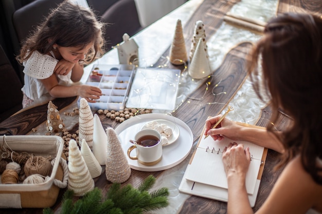 Junge Frau, die Tee trinkt und Pläne oder Ziele für Neujahr 2021 schreibt, während ihre Tochter Weihnachtsbäume aus Papierkegel, Garnen und Knöpfen mit platzierten Sternen und Lichterketten auf Holztisch bastelt.