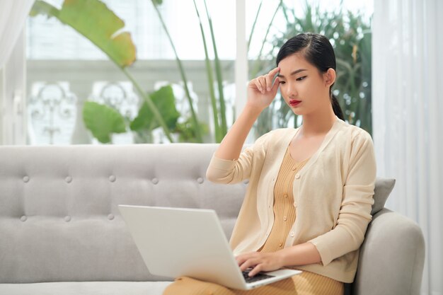 Junge Frau, die unter Kopfschmerzen leidet und wegschaut, während sie den Laptop zu Hause benutzt