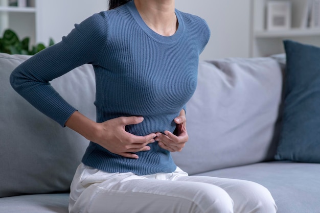 Junge Frau, die unter starken Bauchschmerzen leidet, während sie zu Hause auf dem Sofa sitzt Kranke Frau mit Bauchschmerzen