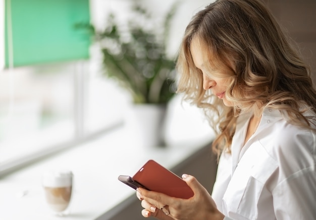 Junge Frau, die Video in den sozialen Netzwerken auf dem Smartphone oder das Senden der Textnachricht während der Pause mit der Kaffeetasse im Geschäftsbüro ansieht