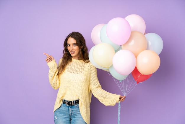 Junge Frau, die viele Luftballons auf lila Wand fängt, die Finger zur Seite zeigt