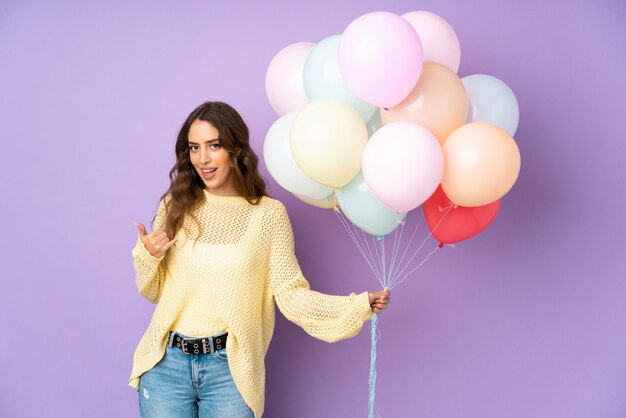 Junge Frau, die viele Luftballons auf lila Wand fängt, die Telefongeste macht