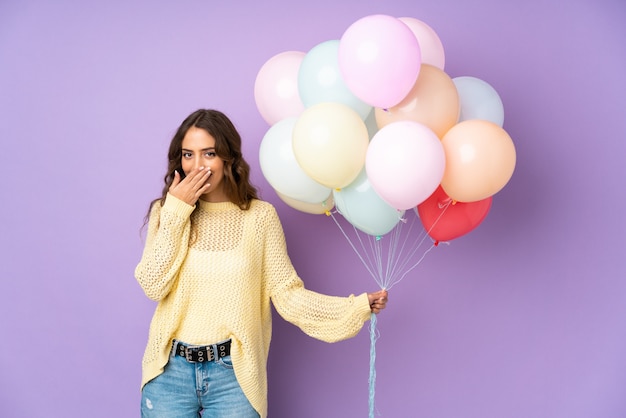 Junge Frau, die viele Luftballons auf lila Wand mit überraschtem Gesichtsausdruck fängt