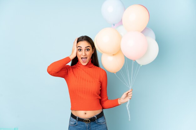 Junge Frau, die viele Luftballons fängt, die auf blaue Wand lokalisiert überrascht und Finger zur Seite zeigen