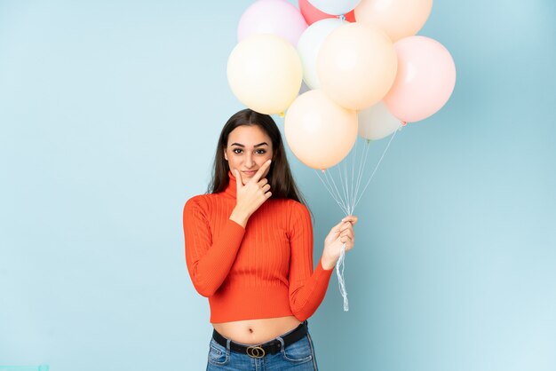 Junge Frau, die viele Luftballons lokalisiert auf blauer Wand fängt, die eine Idee denken