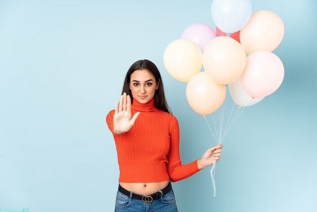 Junge Frau, die viele Luftballons lokalisiert auf blauer Wand fängt, die Stoppgeste mit ihrer Hand macht