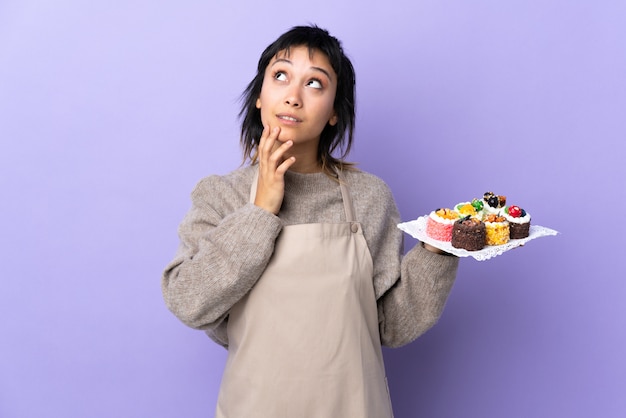 Junge Frau, die viele verschiedene Minikuchen über isoliertem Purpur hält, der eine Idee denkt