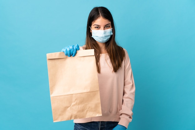 Junge Frau, die vom Coronavirus mit einer Maske schützt und eine Einkaufstüte hält, die auf blauer Wand mit glücklichem Ausdruck lokalisiert wird