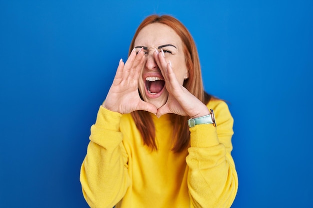 Junge Frau, die vor blauem Hintergrund steht und wütend laut mit den Händen über dem Mund schreit