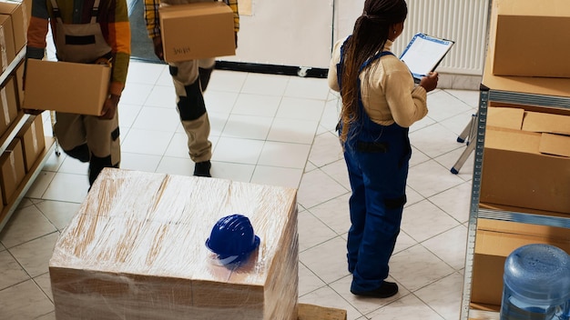 Junge Frau, die vor dem Transport Kisten mit Produkten überprüft und Pakete mit Waren für den Versand vorbereitet. Zwei Männer tragen Kartons auf Paletten zur Qualitätskontrolle, Warenlager.