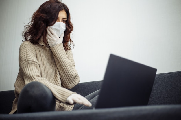 Junge Frau, die während der Quarantäne aufgrund einer Coronavirus-Pandemie von zu Hause aus arbeitet. Schönes Mädchen bleibt zu Hause, trägt medizinische Maske und Handschuhe und tippt auf einem Laptop. Weltweites Konzept der Covid-19-Epidemie.