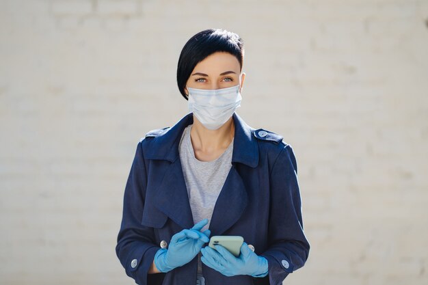 Junge Frau, die während des Ausbruchs von Covid 19 eine medizinische Einwegmaske und Handschuhe mit Smartphone auf der Straße trägt. Schutz in der Prävention des Coronavirus. Ein Konzept der Lieferung, Online-Service, App.